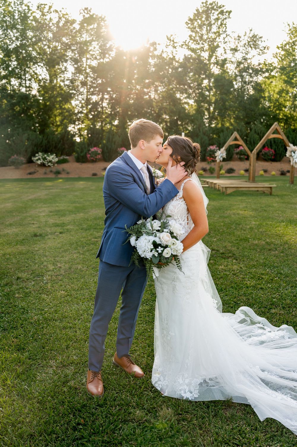 A romantic sunset wedding photo of a couple kissing on a lawn with white floral bouquet.