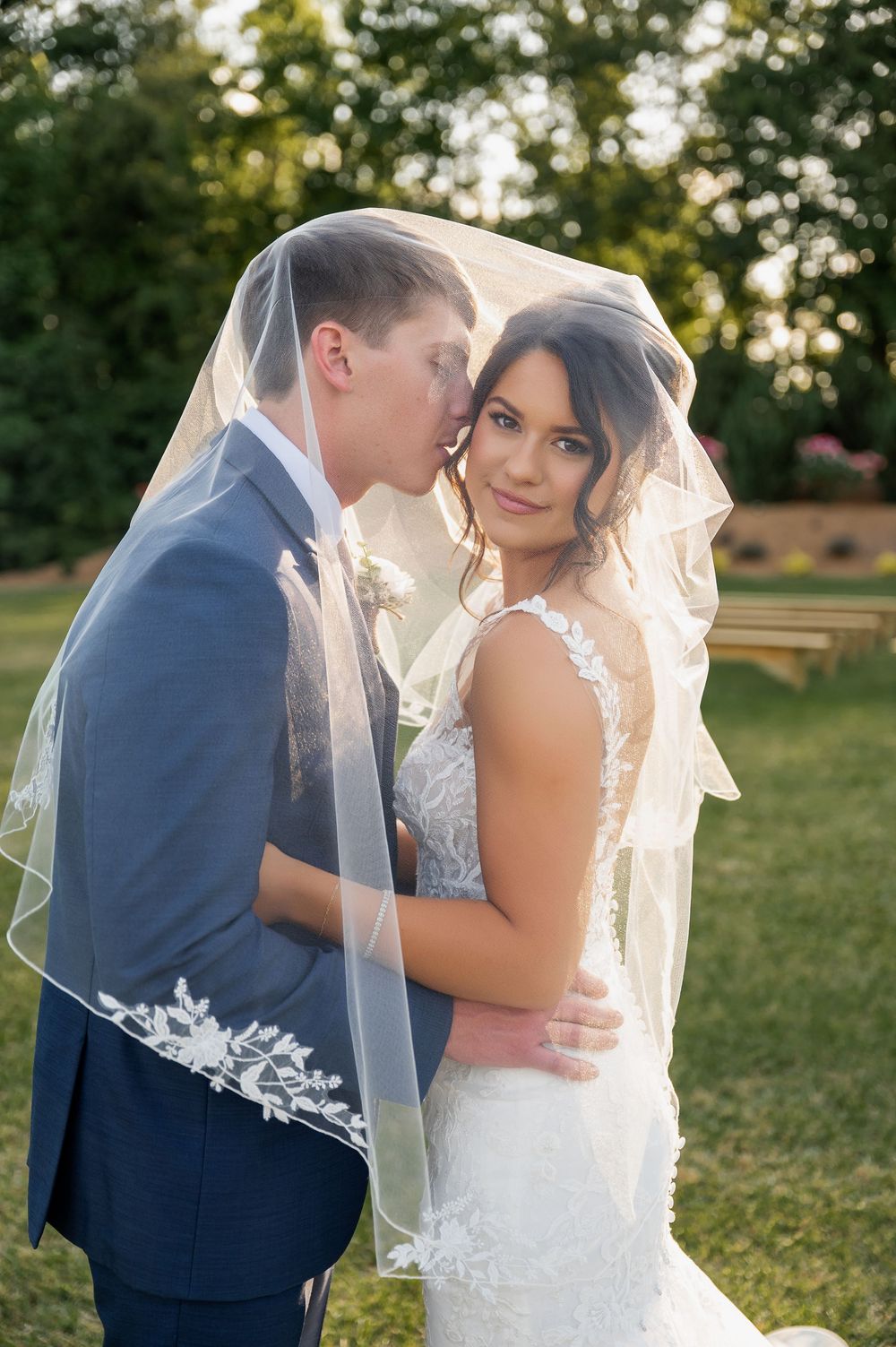 Tender moment between newlyweds sharing a kiss under a flowing veil outdoors.