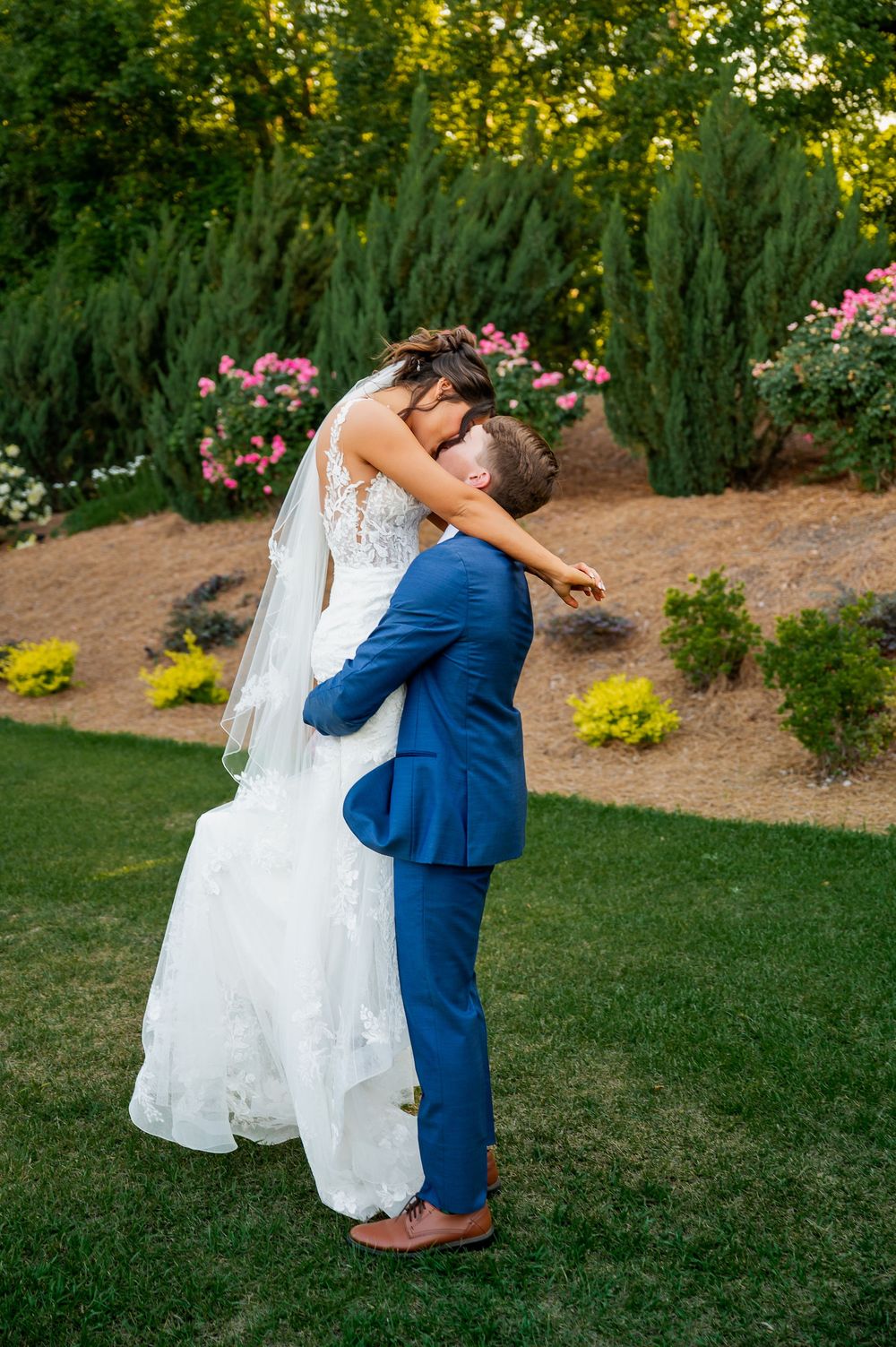 Joyful wedding couple embracing on manicured lawn with colorful garden backdrop.