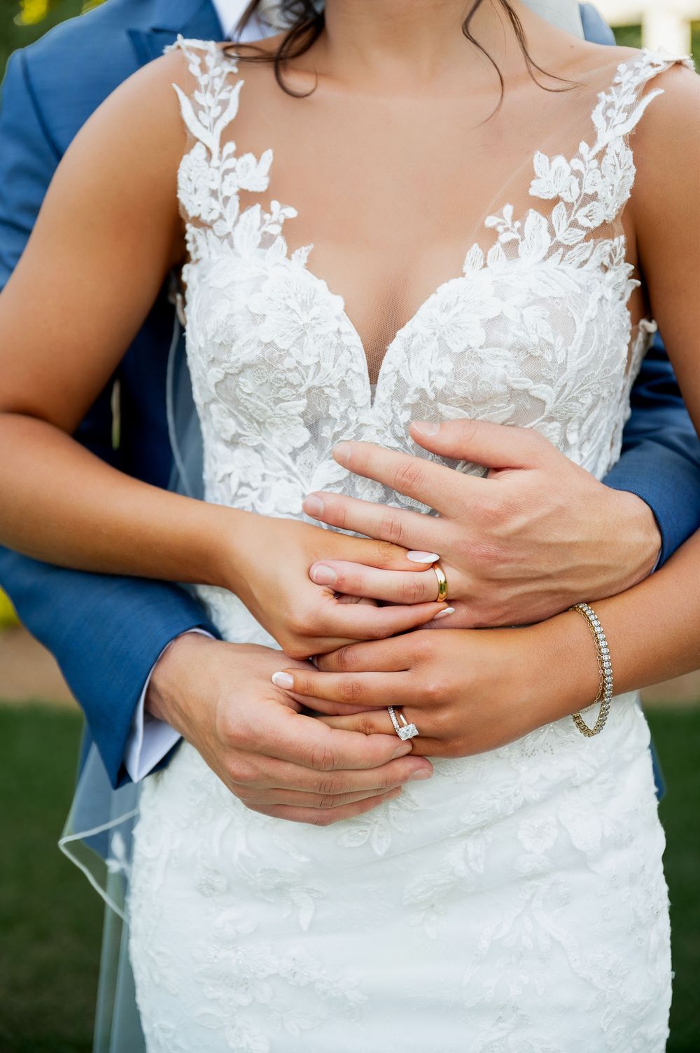 Close-up of hands with wedding rings against detailed lace wedding dress.