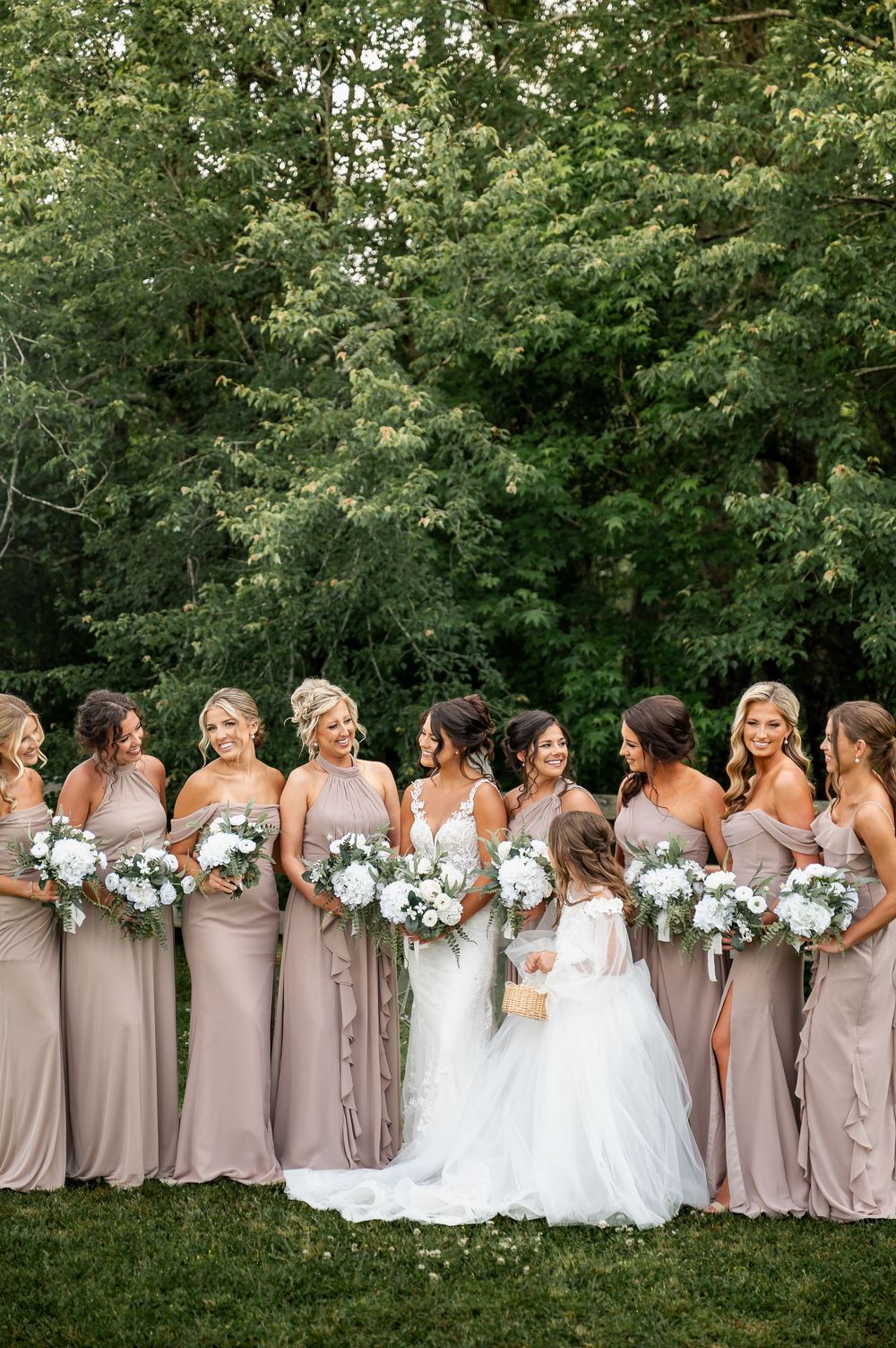 Bridesmaids in matching mauve dresses pose with brides holding white bouquets in outdoor setting.