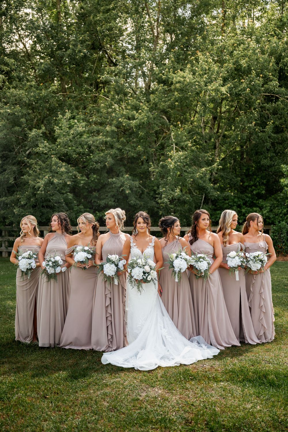 Bridesmaids in taupe dresses holding white bouquets pose together on grassy lawn with trees in background.
