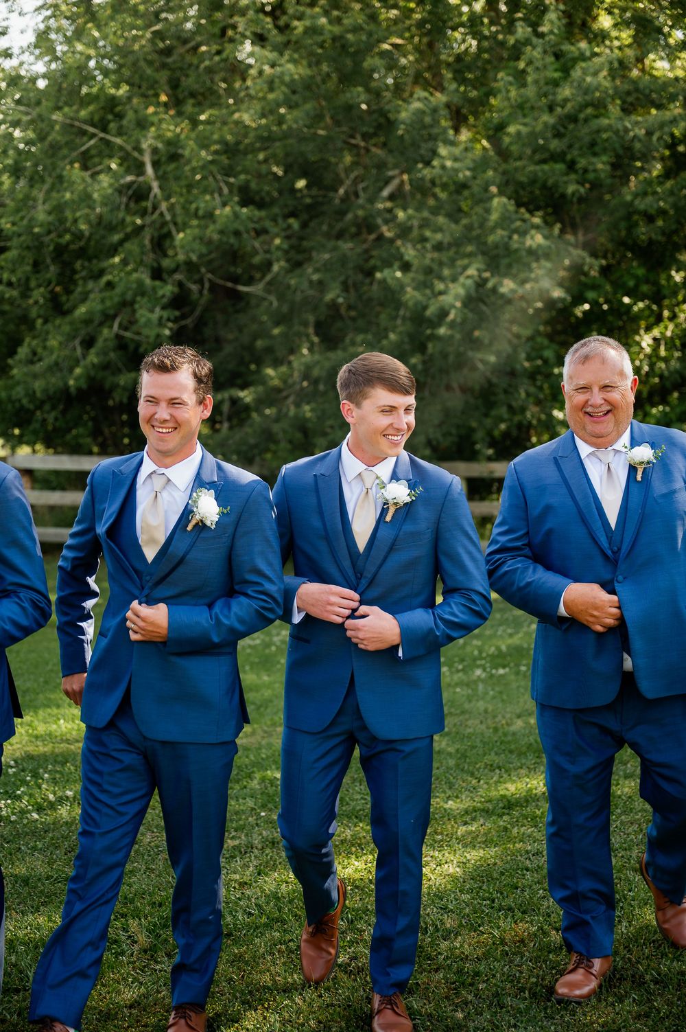 Groomsmen in navy blue suits and brown shoes share laughs while posing on grass.