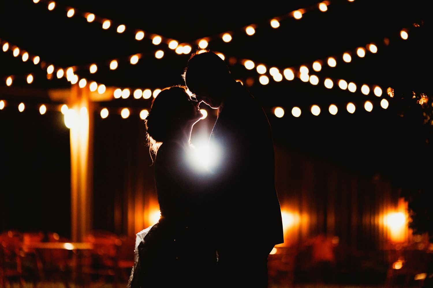 Silhouetted couple sharing an intimate moment under glowing string lights at night.