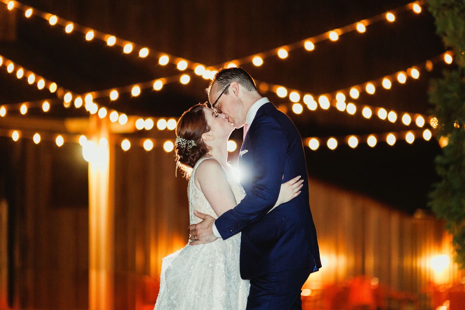Romantic evening portrait under warm string lights at an outdoor wedding celebration.