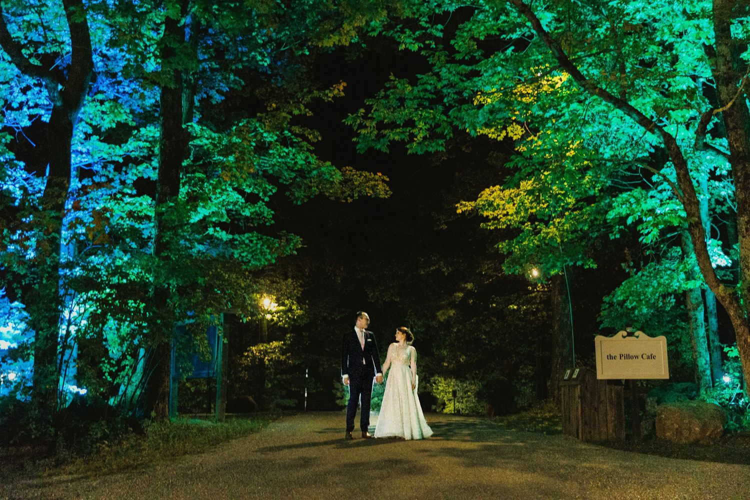 Wedding couple standing on a wooded path illuminated by green uplighting at dusk.
