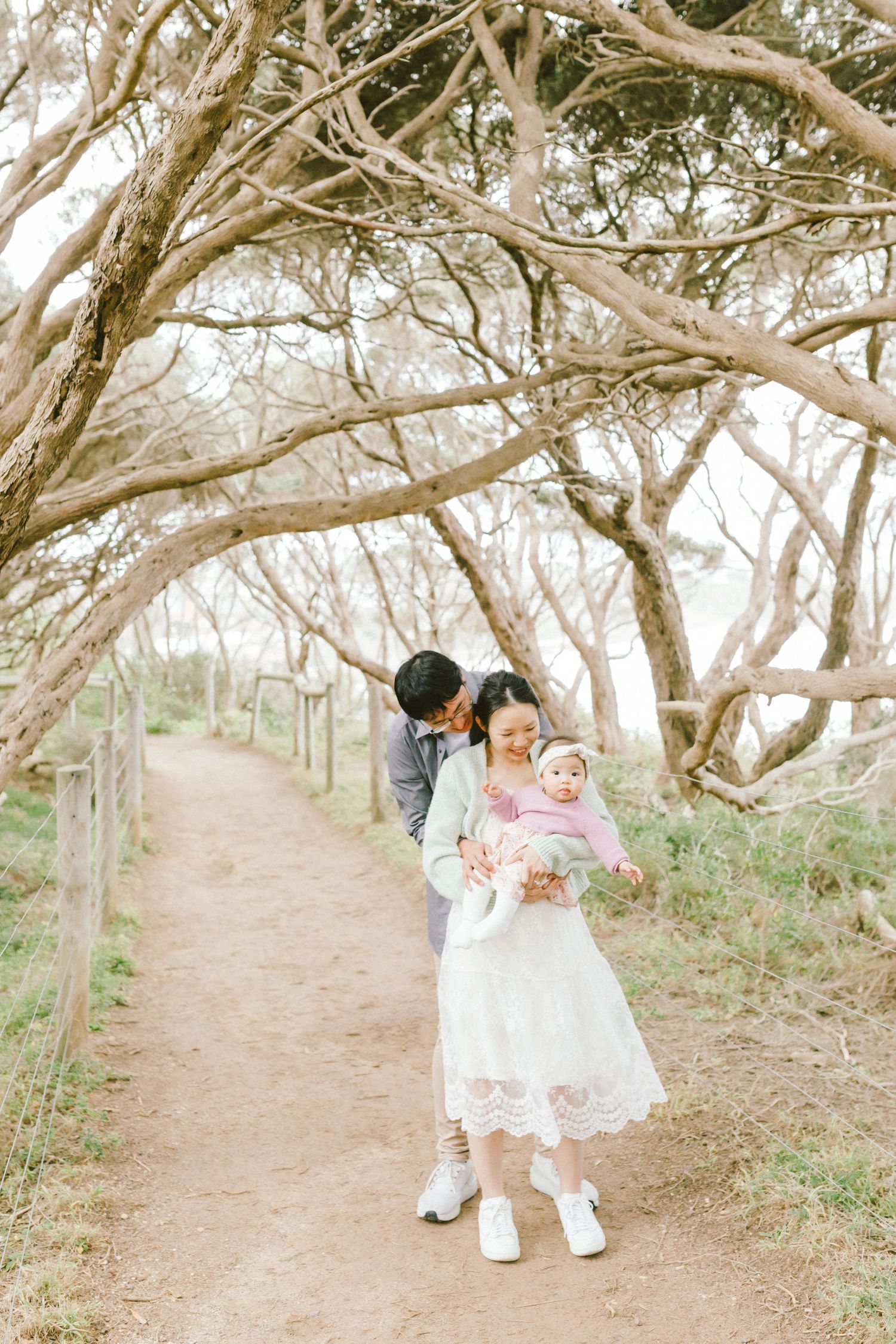 A family walks together through a dreamy wooded path lined with twisted tree branches on a bright sunny day.