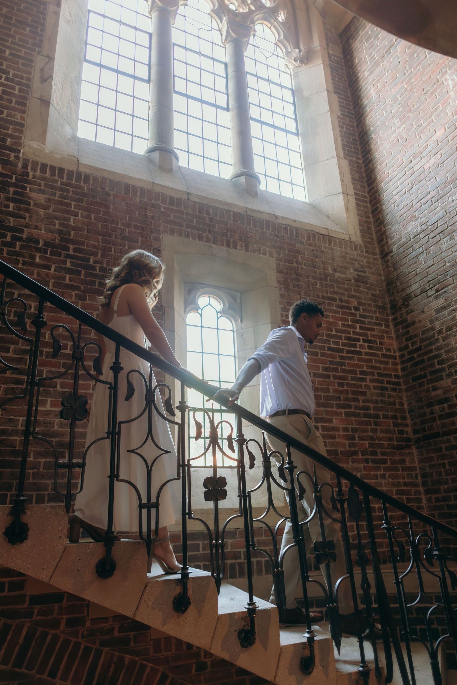 Sunlight streams through Gothic windows onto a curved staircase with ornate black iron railings in a historic brick building.