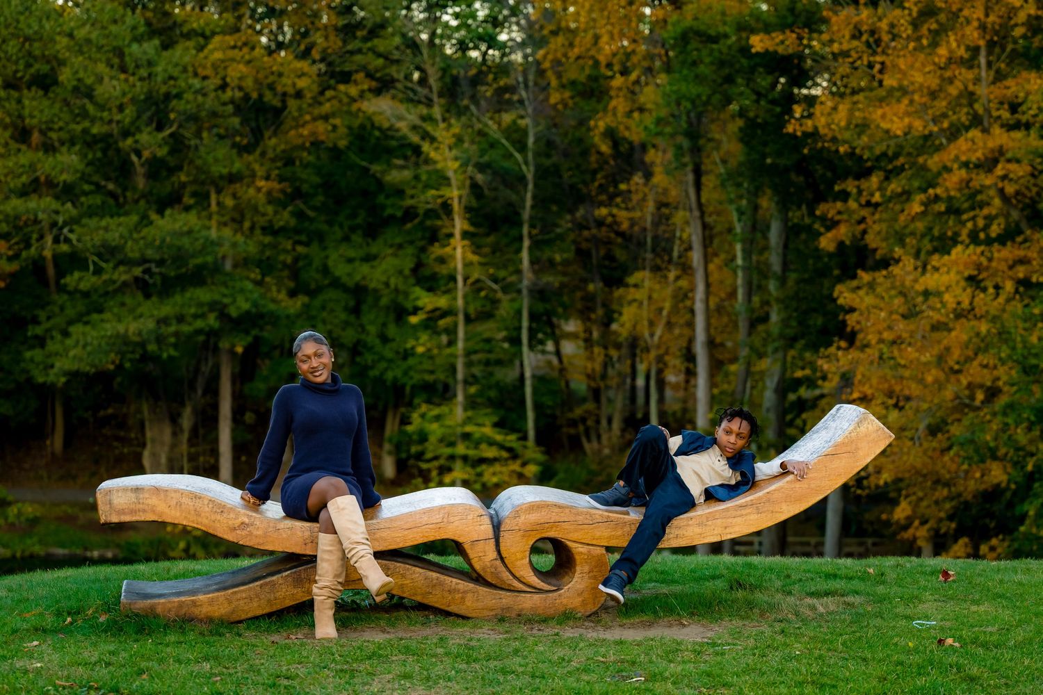 A wooden sculpture installation in a park setting shows curved organic forms against an autumn forest backdrop.