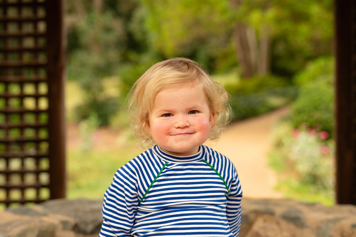A toddler in a navy striped shirt smiles while sitting outdoors during a family photo session in Northern Virginia; Arlington Virginia.