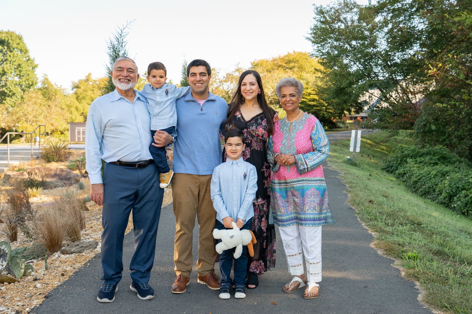 A family of four plus grandparents poses on a paved path surrounded by autumn foliage and greenery during a family photography session.