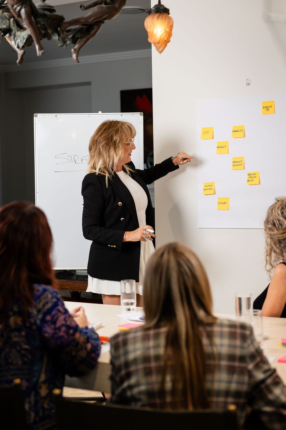 A presenter in black attaches sticky notes to a whiteboard during a workshop session with attendees seated in rows.
