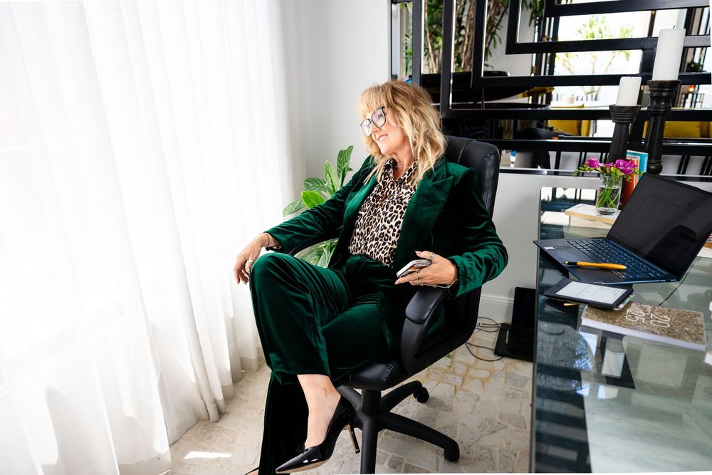 Professional office worker in emerald green blazer sits at modern desk with laptop and paperwork.
