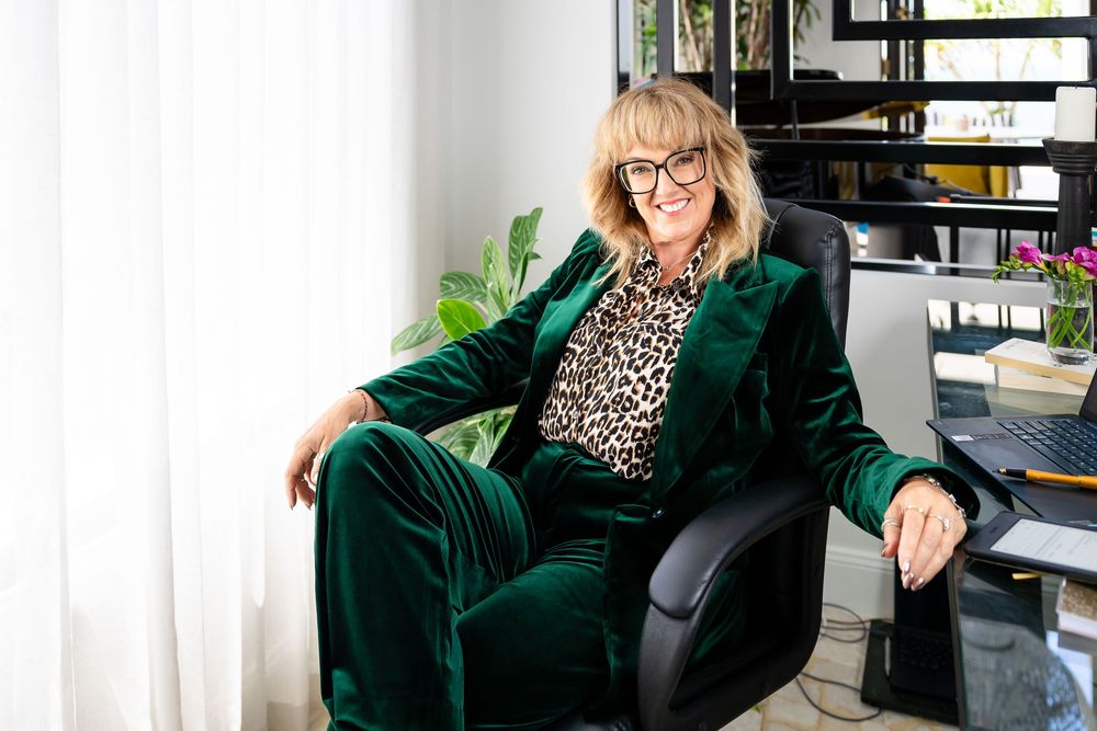 A person lounges on an emerald green velvet sofa wearing a leopard print top and dark blazer in a modern living space.