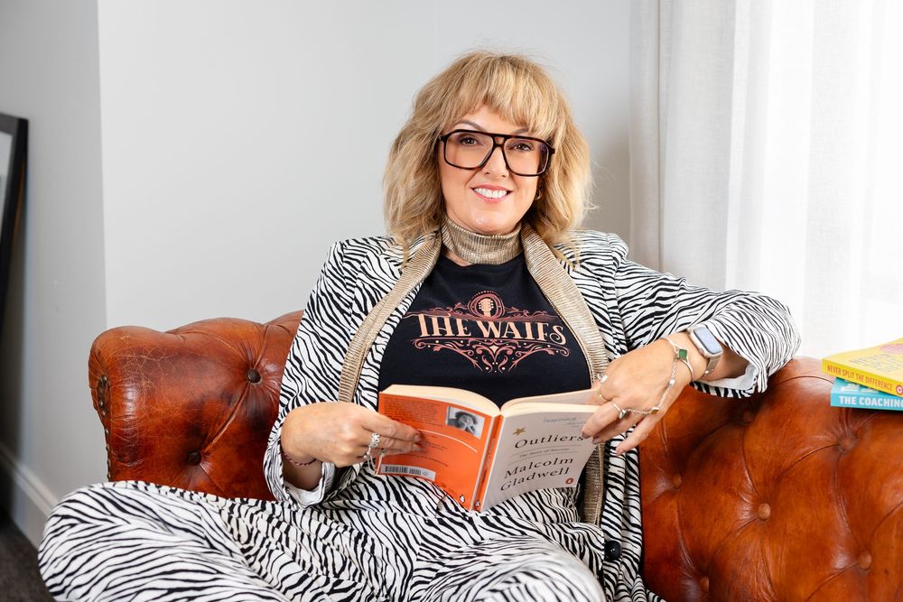 A person wearing glasses sits comfortably on a brown leather couch while reading a book.