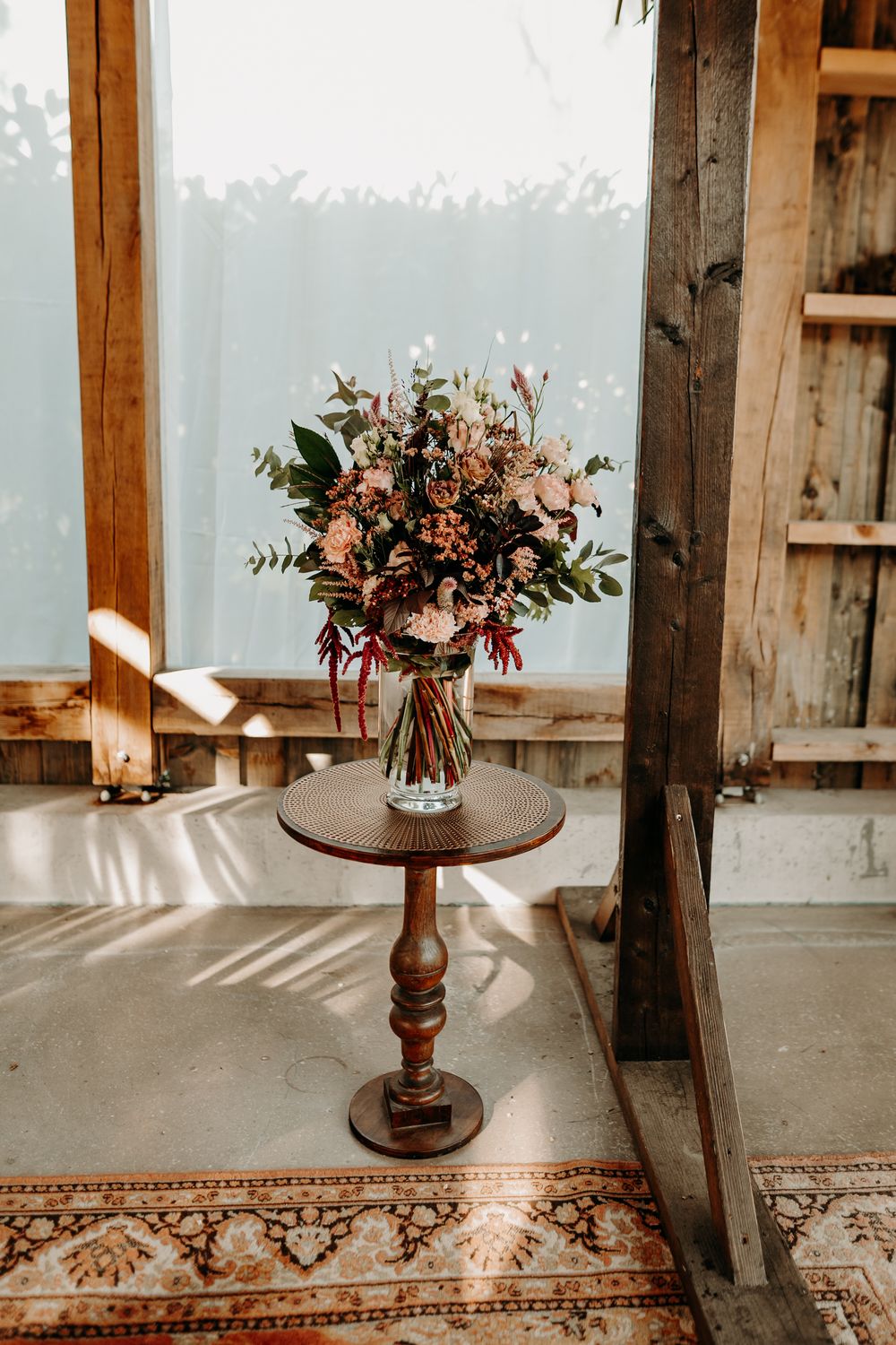 Rustic wedding decor features floral arrangements on wooden pedestals with an ornate rug in a sunlit barn venue.
