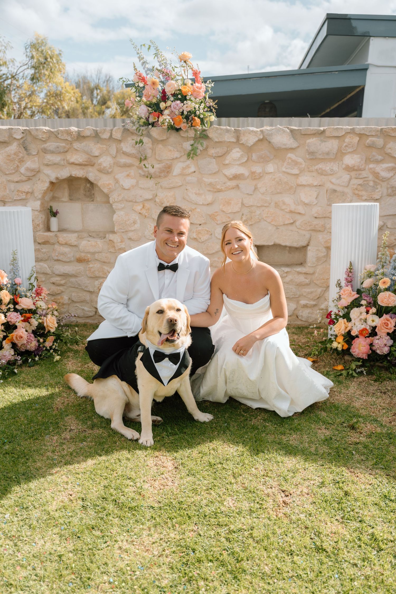 Wedding couple sits on grass with their adorable golden retriever wearing a tuxedo bow tie beside stone wall.