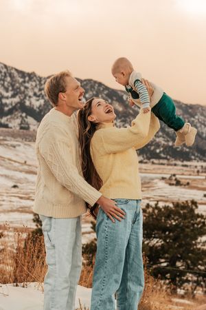 parents in cream and yellow sweaters lifting their baby outdoors in the snowy mountains  | Motherhood + Family Photographer Westminster, CO