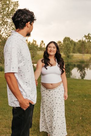 Pregnant woman in white crop top and floral skirt smiling at partner outdoors near a pond. Motherhood + Family Photographer Westminster, C0