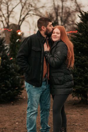 A couple shares an intimate moment among evergreen trees at dusk during the holidays. | Motherhood + Family Photographer Westminster, CO
