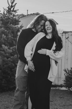smiling pregnant couple embracing outdoors near evergreen trees | Motherhood + Family Photographer Westminster, CO