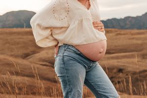 Pregnant woman in white blouse + jeans cradling baby bump in golden field with mountains. | Motherhood + Family Photographer Westminster, CO