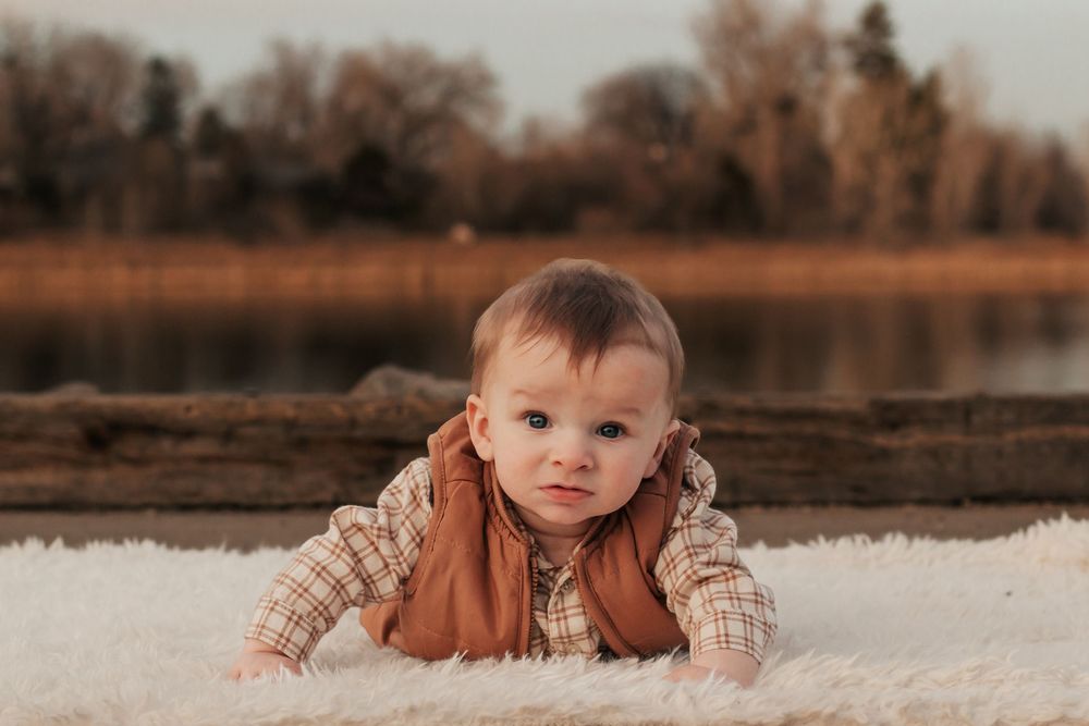 A baby in warm clothing sits outdoors in autumn with a lake and trees in the background. | Motherhood + Family Photographer Westminster, CO