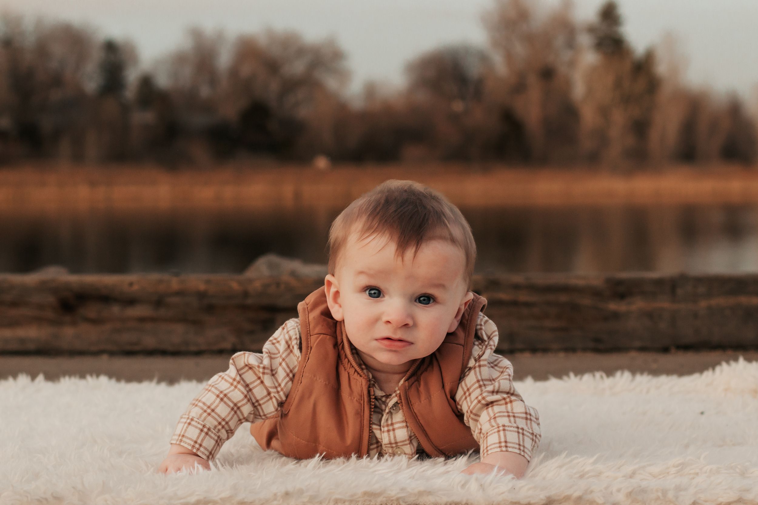 A baby in warm clothing sits outdoors in autumn with a lake and trees in the background. | Motherhood + Family Photographer Westminster, CO