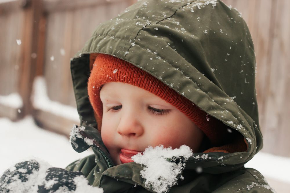 A child wearing a green winter hat catches snowflakes with their tongue during a snowy day. | Motherhood + Family Photographer Westminster,