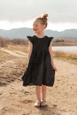 A young child wearing a black dress twirls and plays on a dirt path with mountains  | Motherhood + Family Photographer Westminster, CO