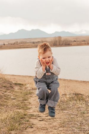 A child in denim overalls jumps playfully by a lake with mountains in the background | Motherhood + Family Photographer Westminster, CO