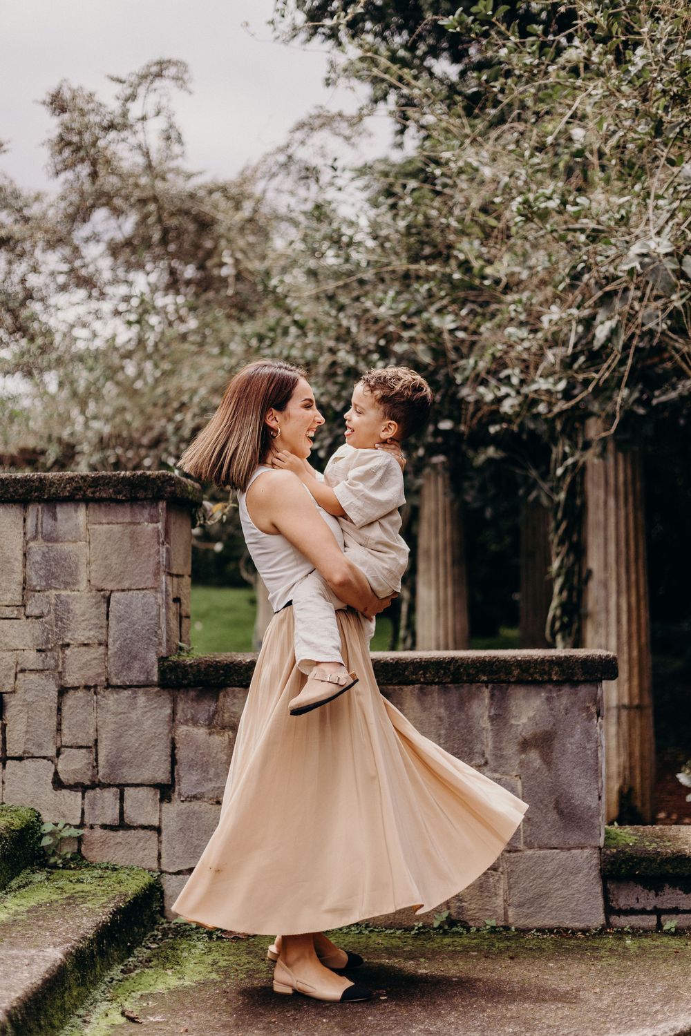 A figure in a flowing beige skirt twirls with a small child in an outdoor garden setting with stone walls.