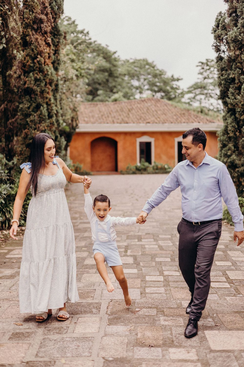 Family walking and playing together on stone pathway in front of rustic orange stucco building with cypress trees.