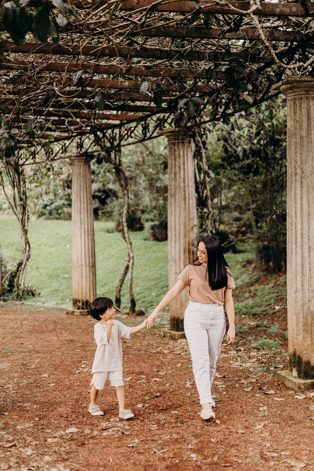 A parent and child walking together under a beautiful pergola covered with vines in a garden setting.