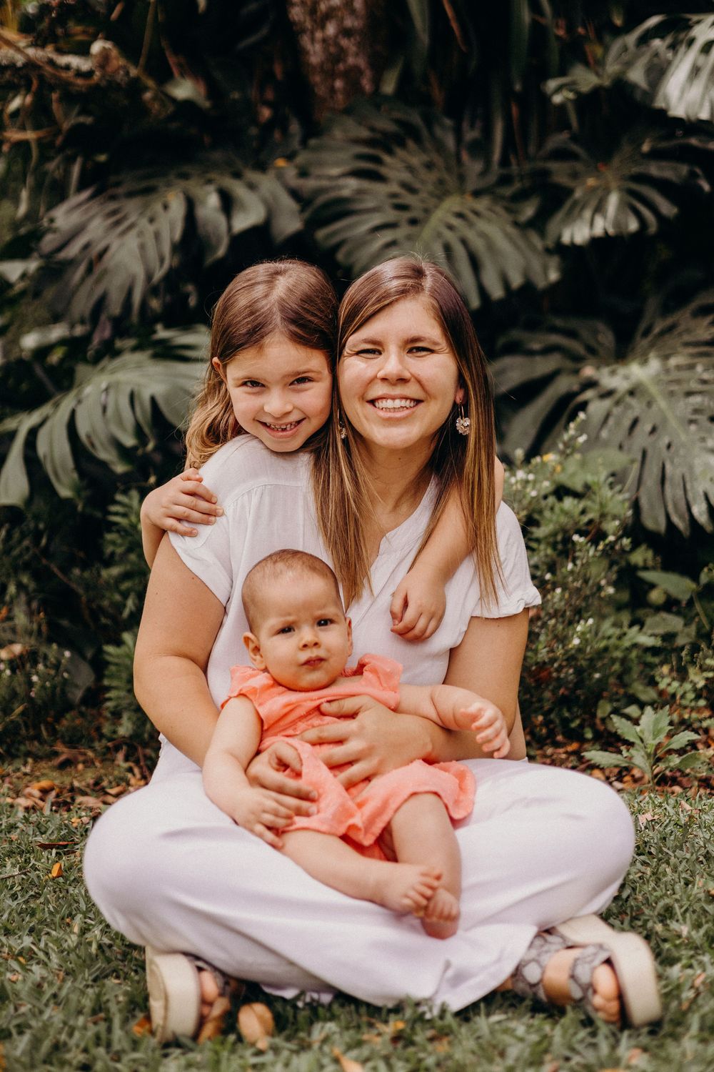 Three people wearing white outfits sit together outdoors among tropical foliage in a casual family portrait setting.