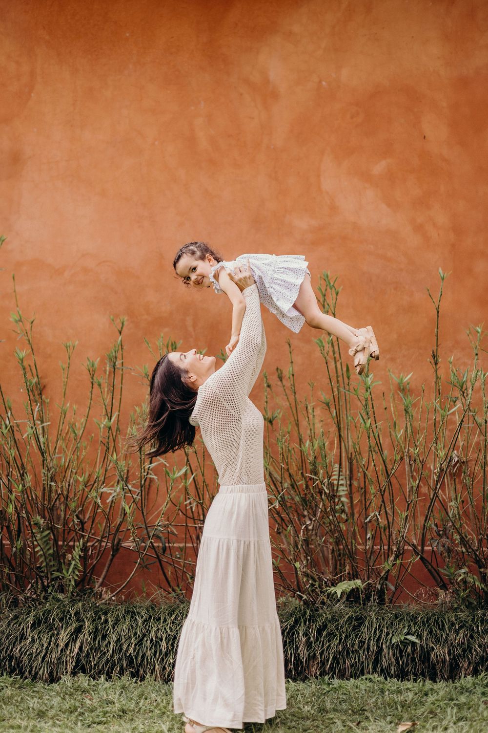 A figure in a flowing white dress performs an expressive dance sequence against a rustic terracotta wall and green foliage.