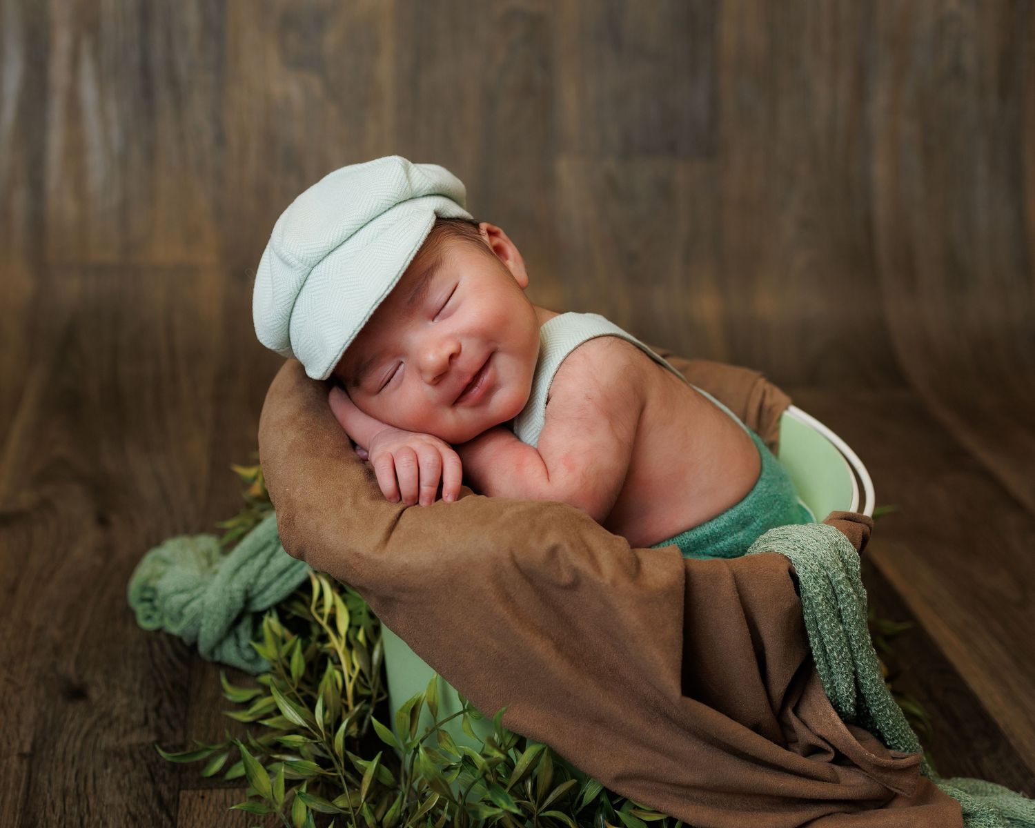 Newborn photography featuring a sleeping baby wrapped in a cozy brown bear-themed knit blanket with greenery accents.