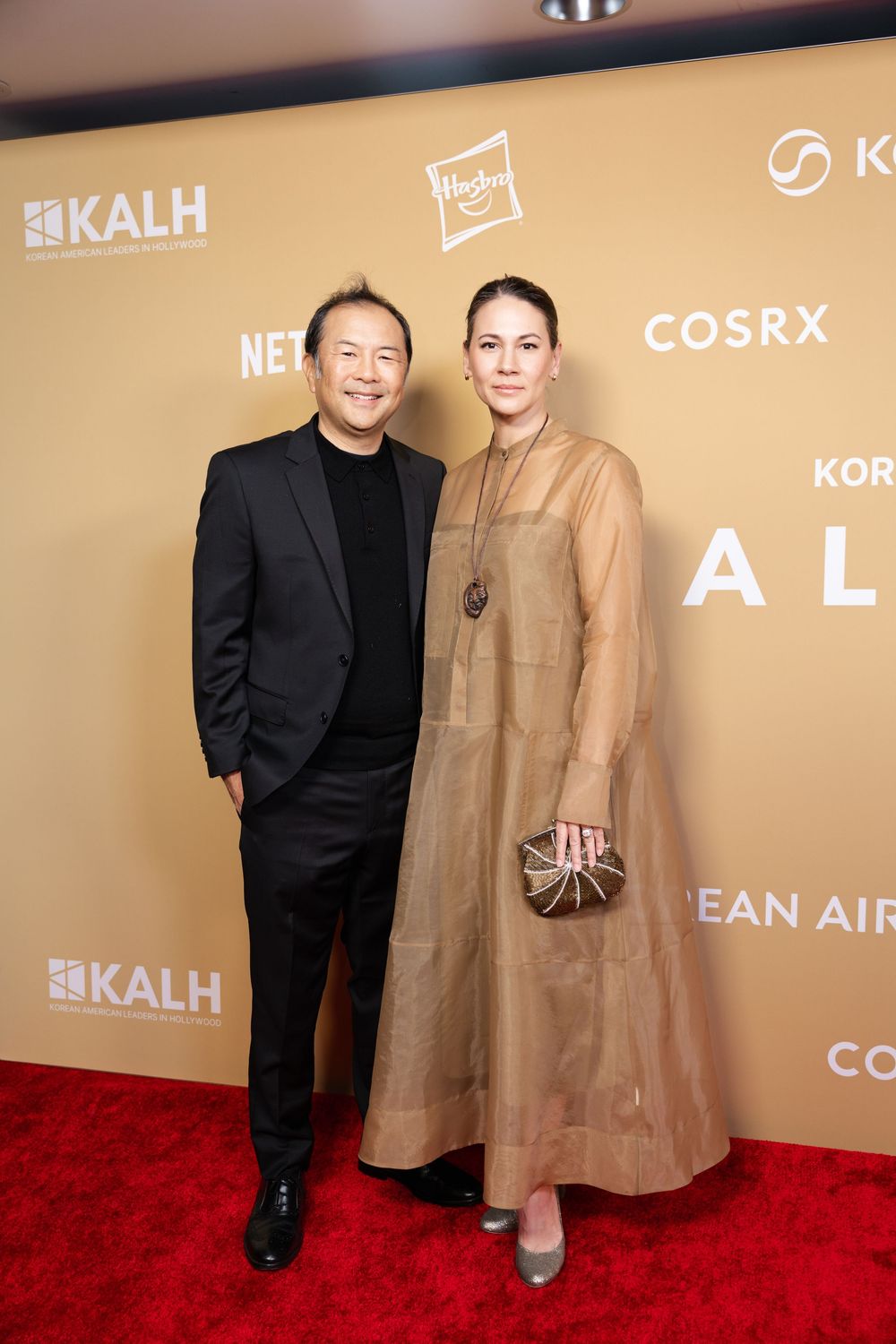 A man in a black suit and a woman in a sheer beige gown pose together on a red carpet event.