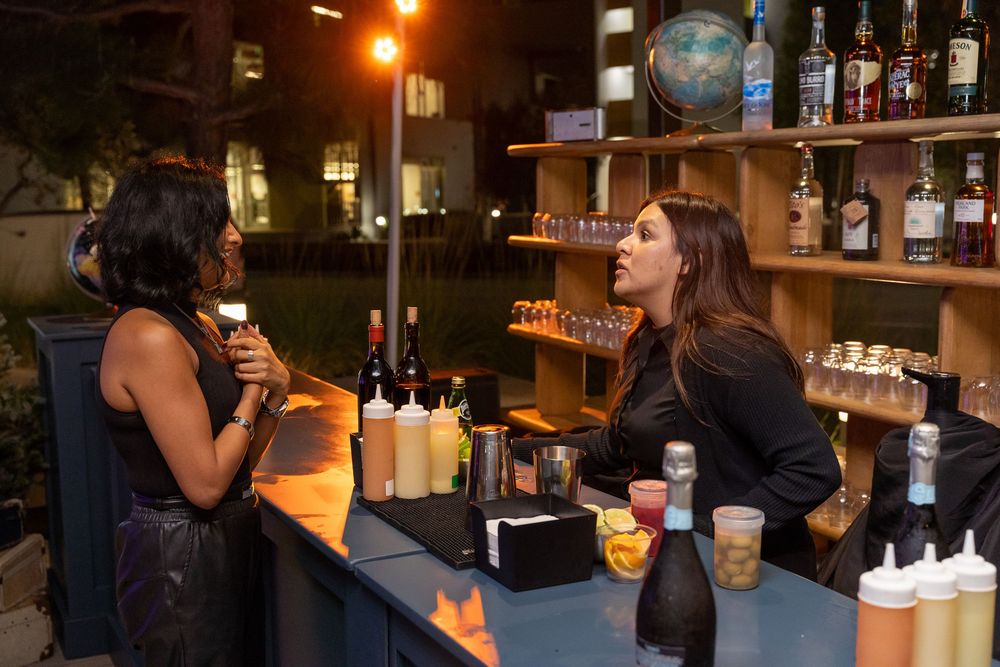 Two women chat at an outdoor bar stocked with bottles and condiments during an evening event.