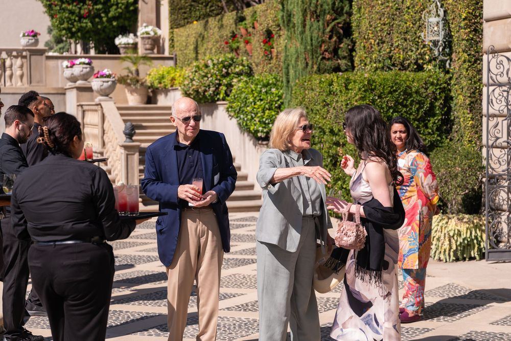 Elegantly dressed guests mingle outdoors at a sophisticated garden party near a classic stone building.