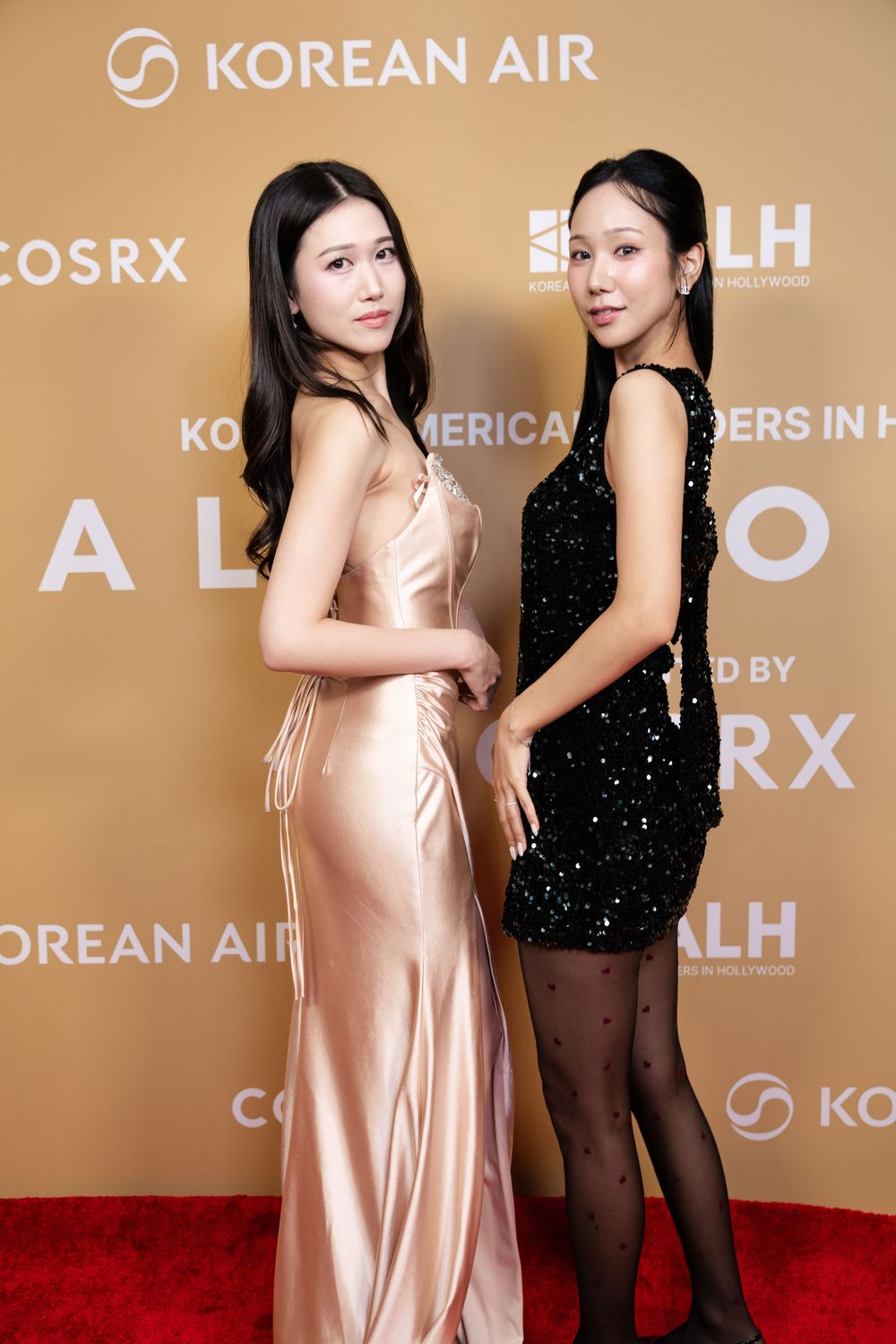 Two women pose on red carpet at Korean Air event, one wearing pink silk gown and other in black sequined dress.