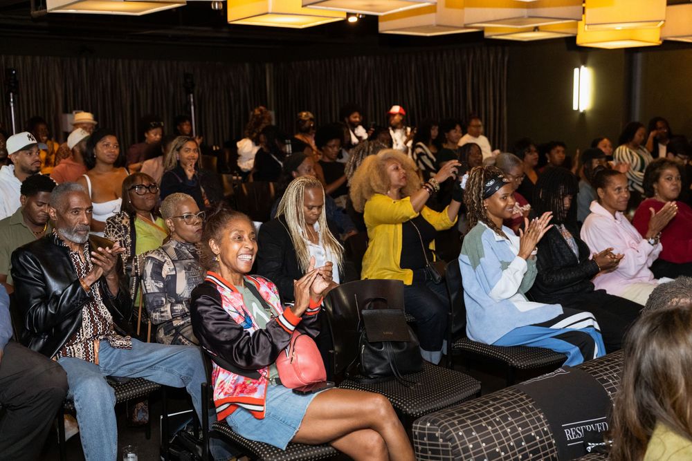 Audience members sit in rows of chairs during an indoor event in a dimly lit venue.