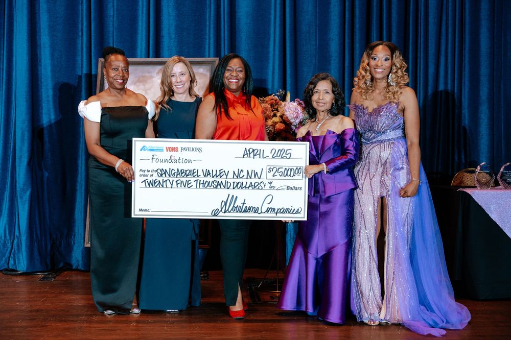A group stands on stage in formal evening wear holding signs during an awards ceremony against blue curtains.