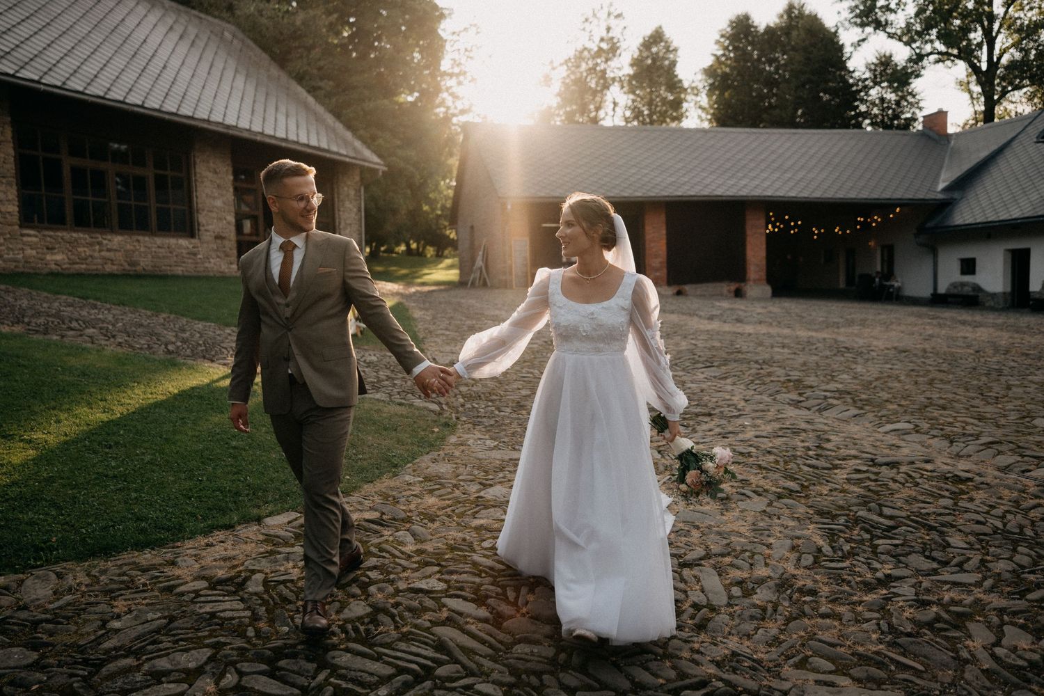 A romantic couple walks hand in hand during sunset at a rustic outdoor venue with golden sunlight filtering through trees.