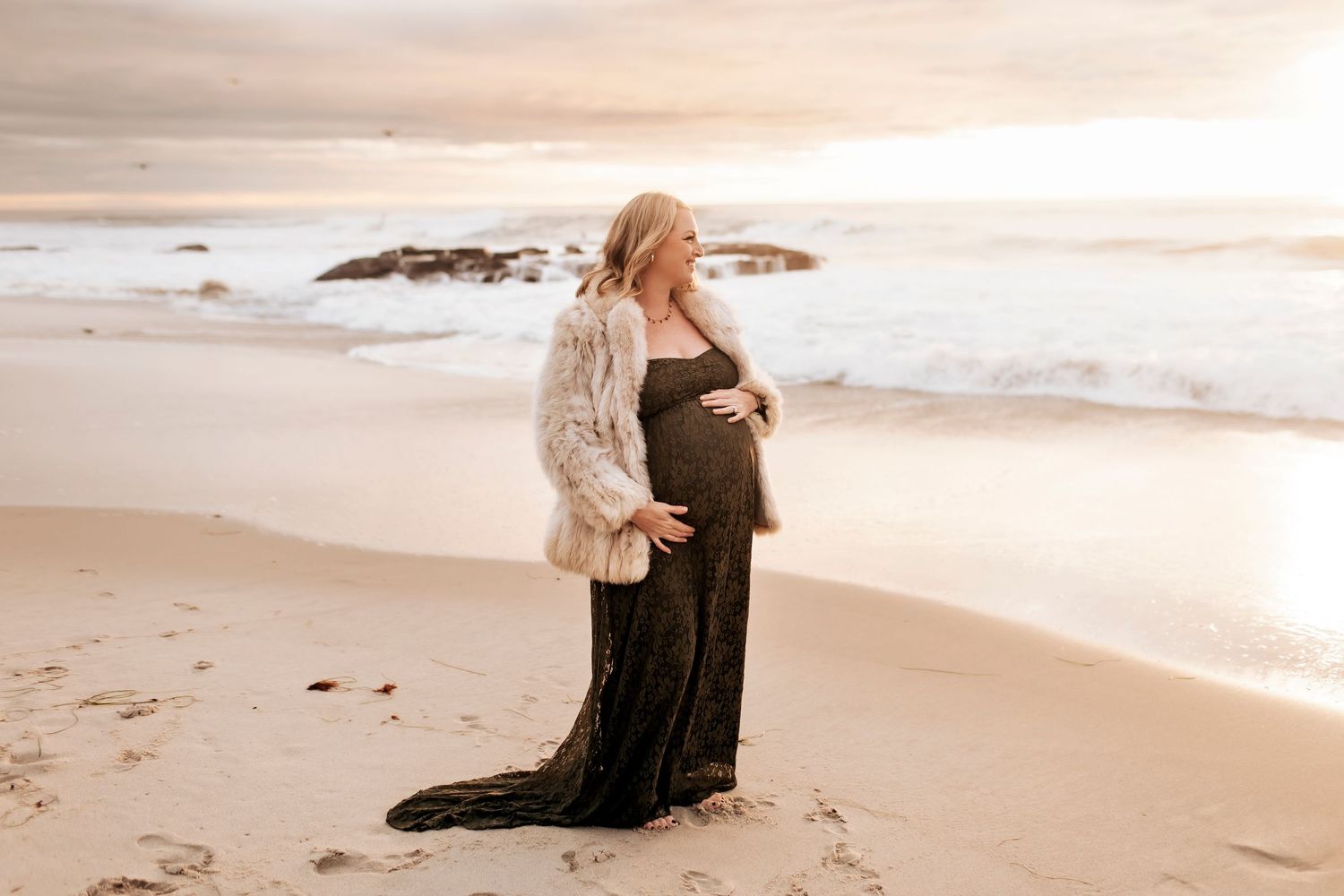 A pregnant woman stands holding her belly looking out at the horizon on a beautiful beach at golden hour.