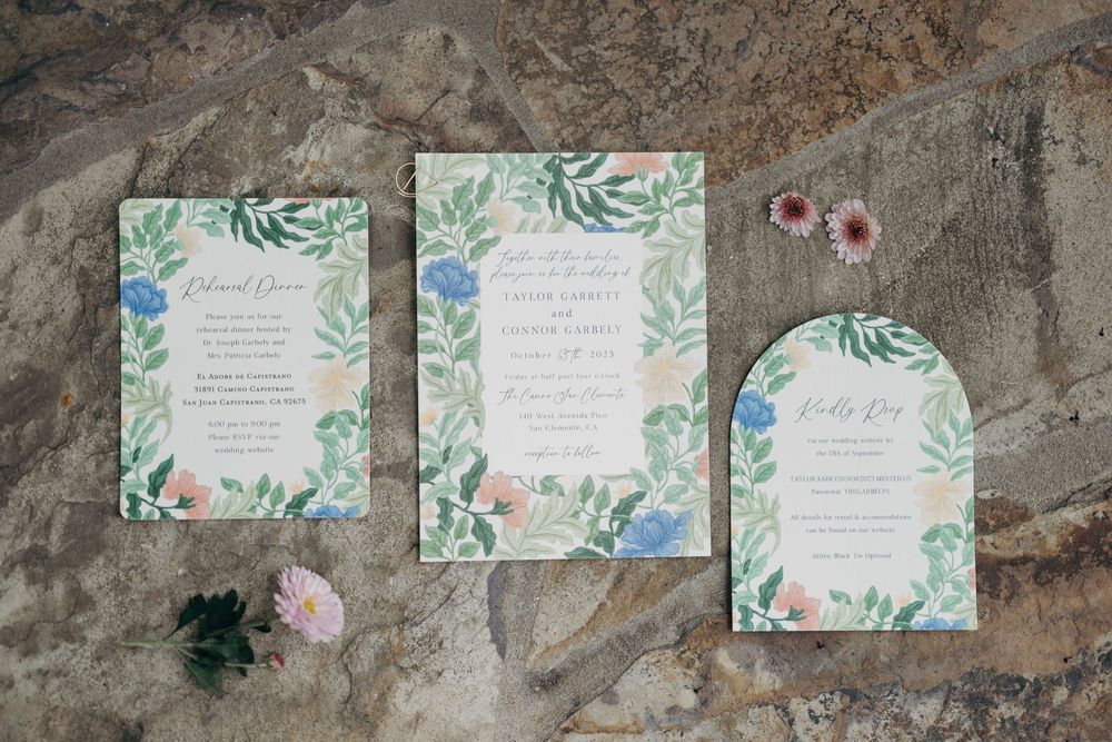 Wedding invitations with watercolor floral designs and greenery displayed on stone surface.