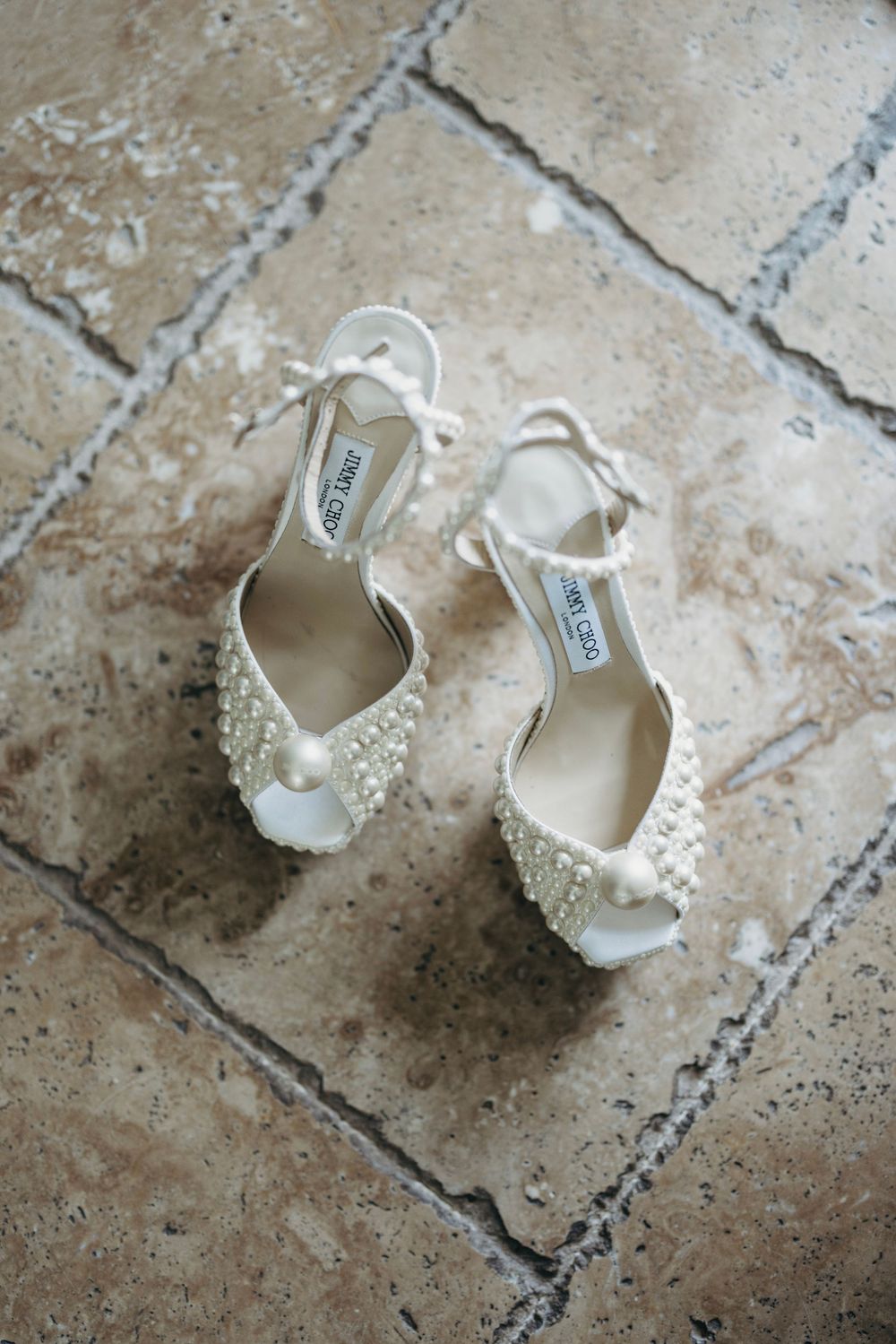 Elegant white bridal shoes with pearl and crystal embellishments on textured stone floor.