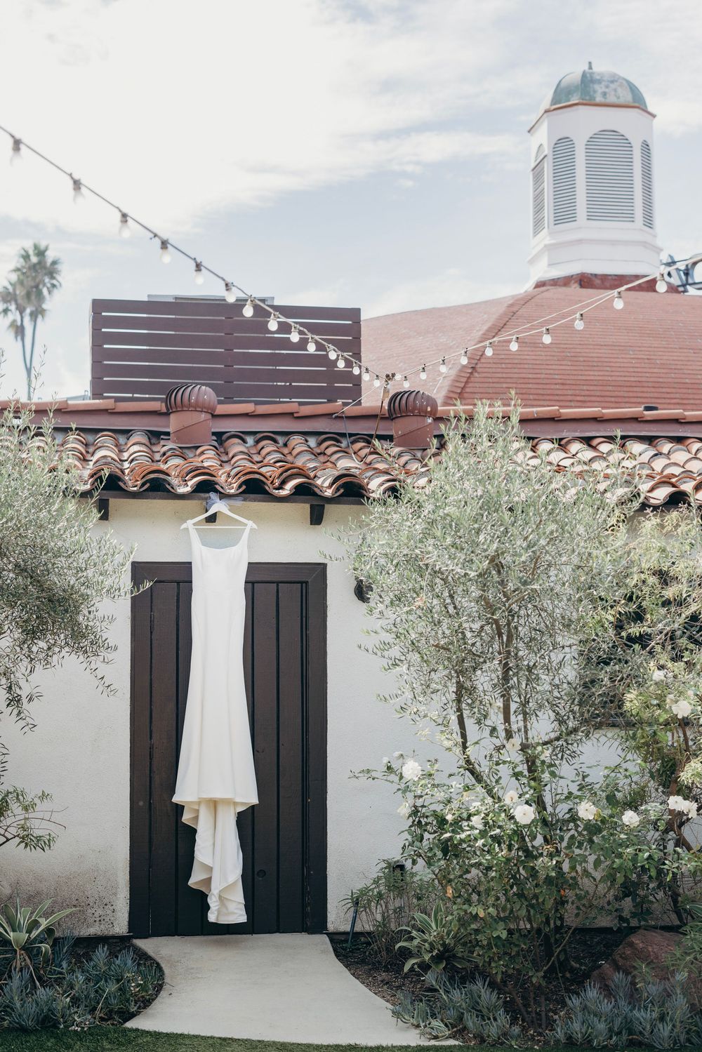 White wedding dress hanging on display against Spanish-style white stucco building with string lights.