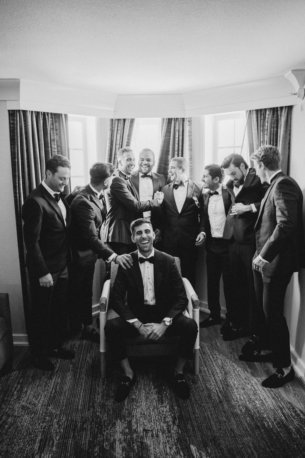 Black and white photo of groomsmen socializing in formal attire at wedding venue.