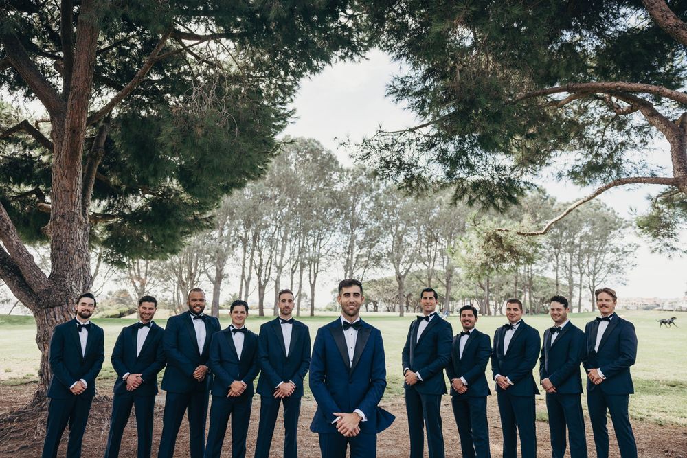 Groomsmen in navy suits pose together outdoors with trees in the background.